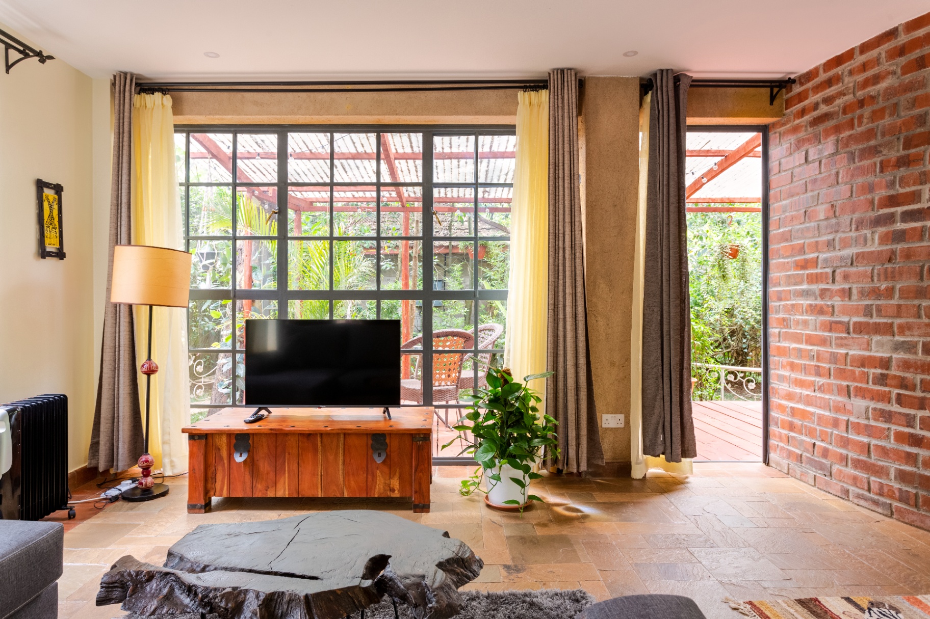 Lounge with timber TV chest, floor-to-ceiling steel-framed windows opening onto a covered garden terrace and brick feature wall