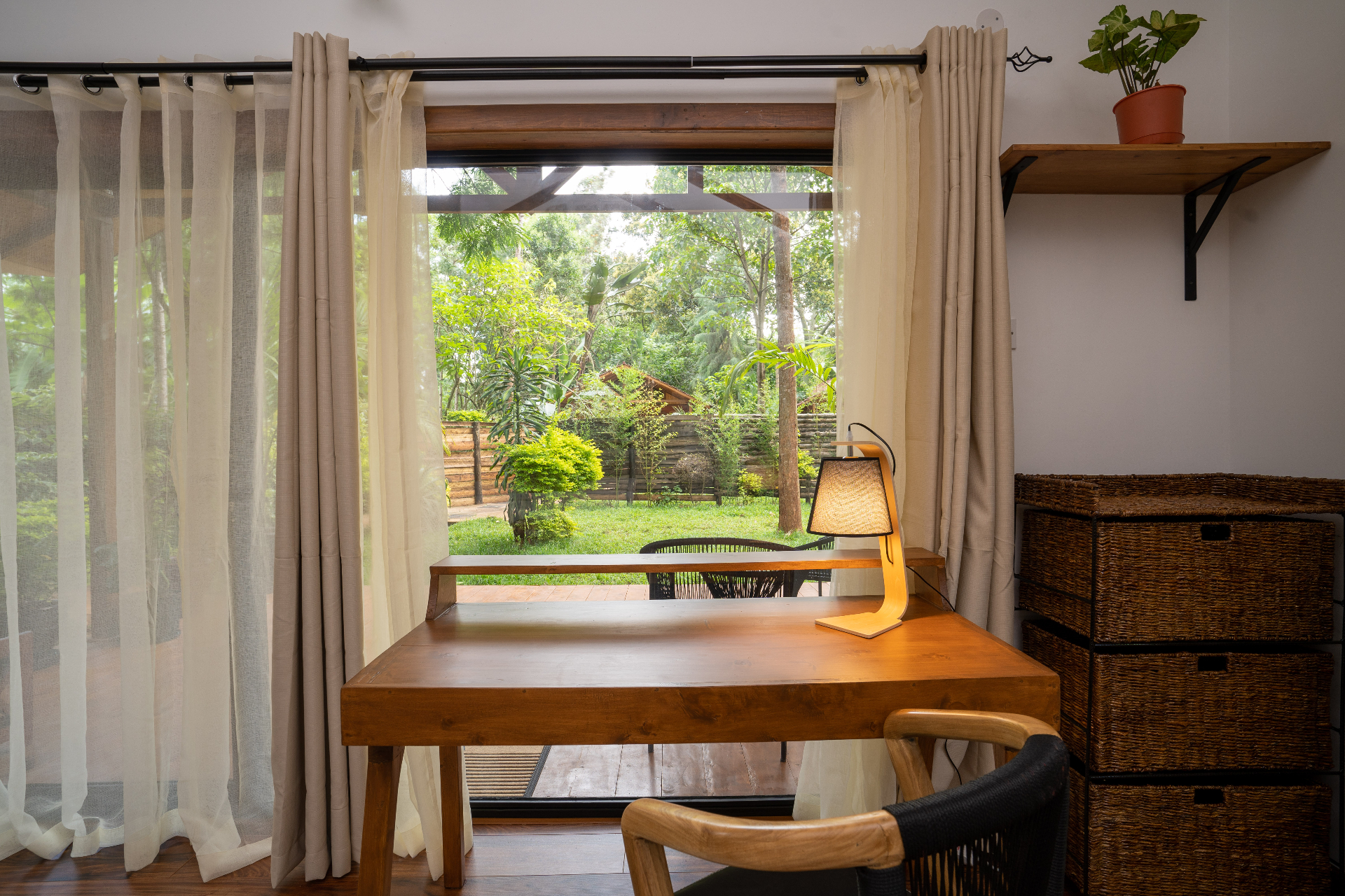 Timber writing desk in front of open glass doors in Cottage 2, looking out to a small private lawn, patio seating and leafy tropical garden beyond