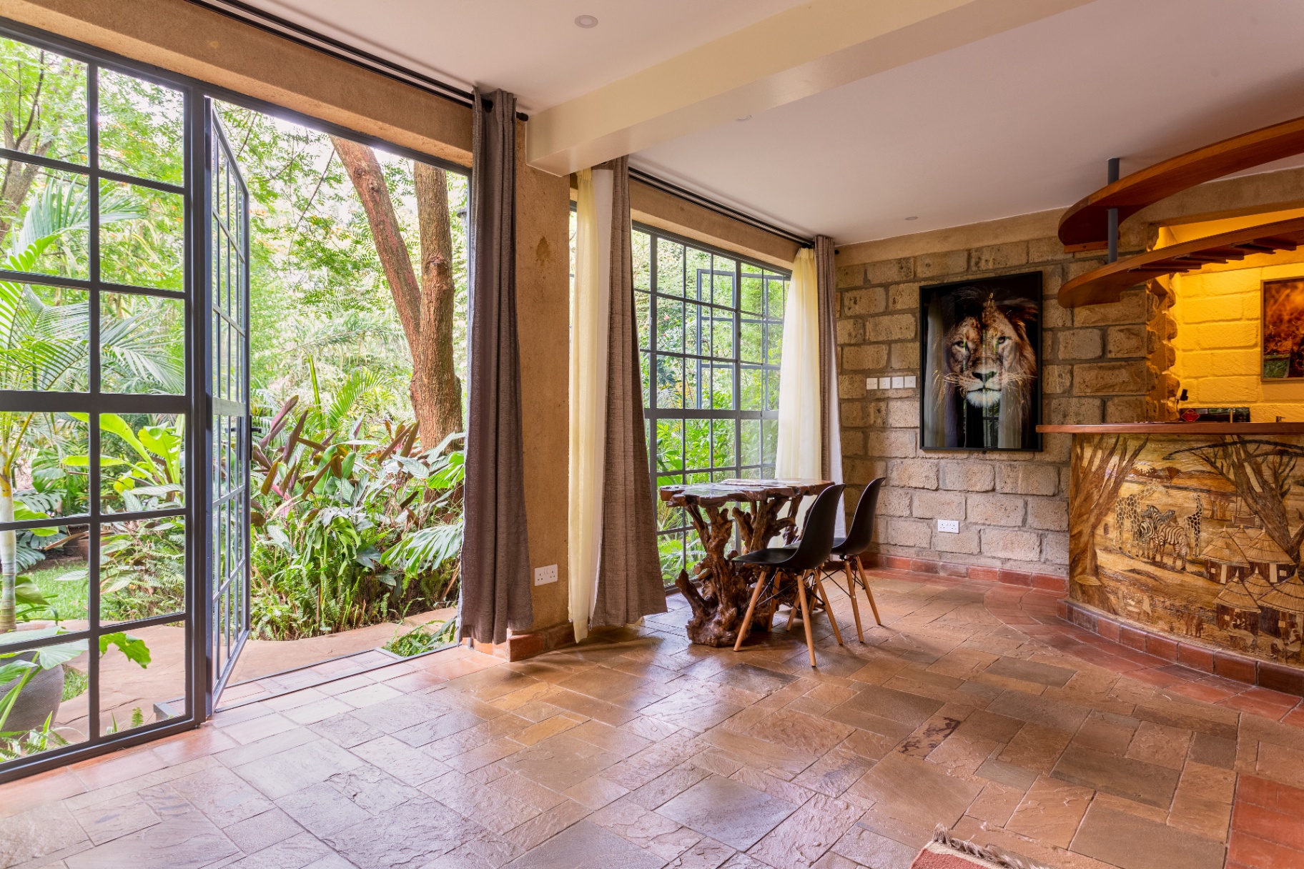 Root-carved dining table with two black chairs beside open steel-framed doors leading to a lush garden, with the curved timber bar and lion artwork to the right