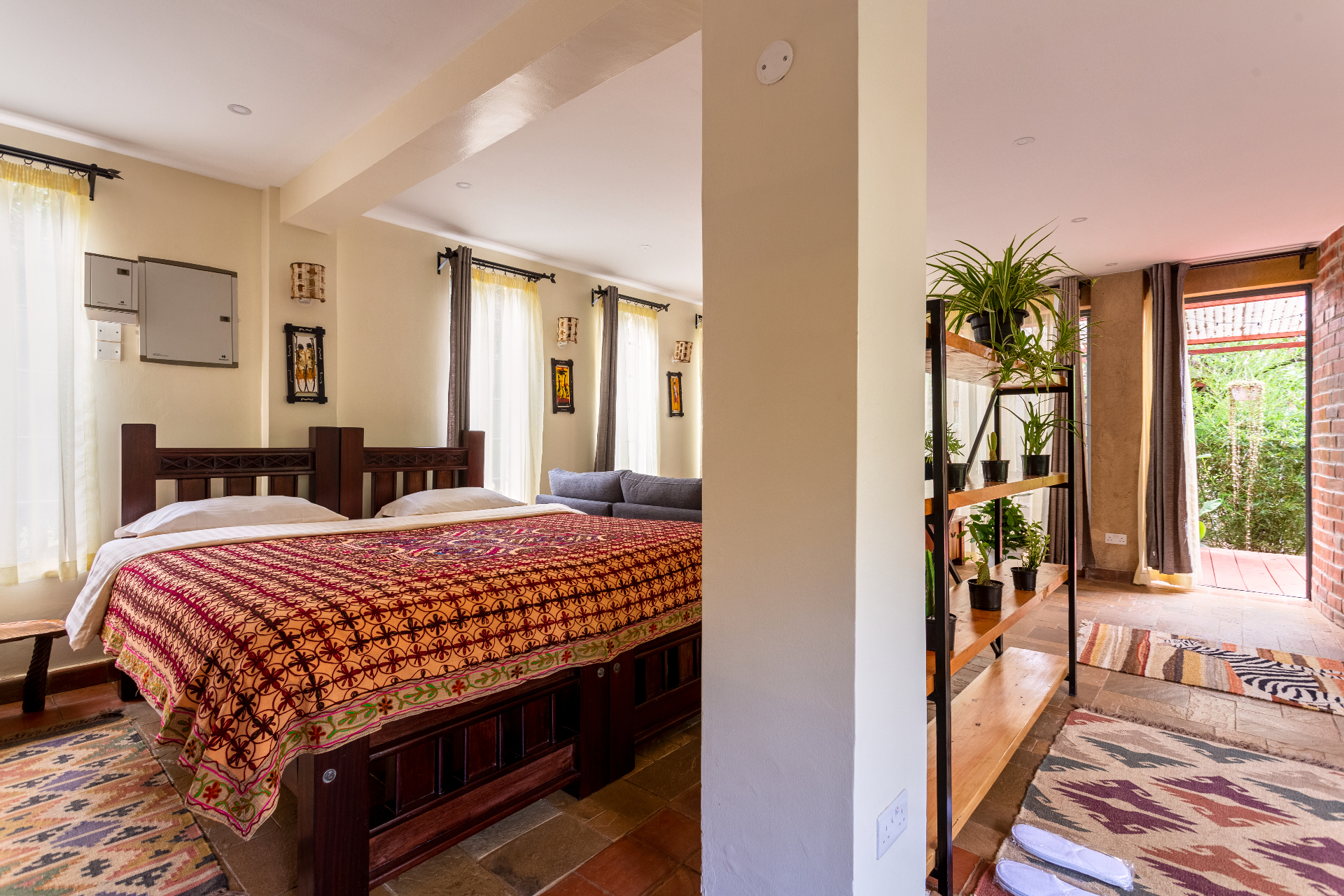 Twin carved-timber beds joined together with an embroidered throw, framed by tall curtained windows and a plant-shelf divider