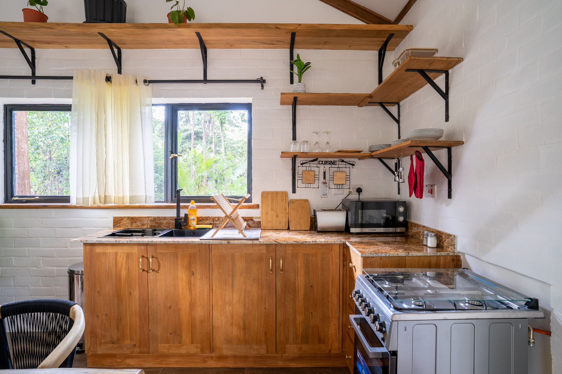 Cottage 2 kitchen counter with granite worktop, black tap, sink, chopping boards, toaster and microwave beside a stainless gas range under open timber shelves