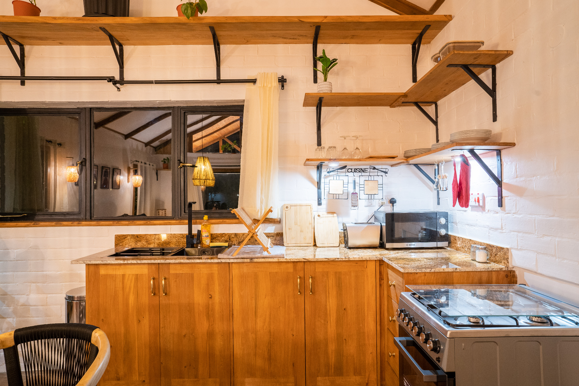 Nighttime view of the Cottage 2 kitchen counter with warm pendant lighting, granite worktops, open shelves, sink, toaster, microwave and stainless gas range