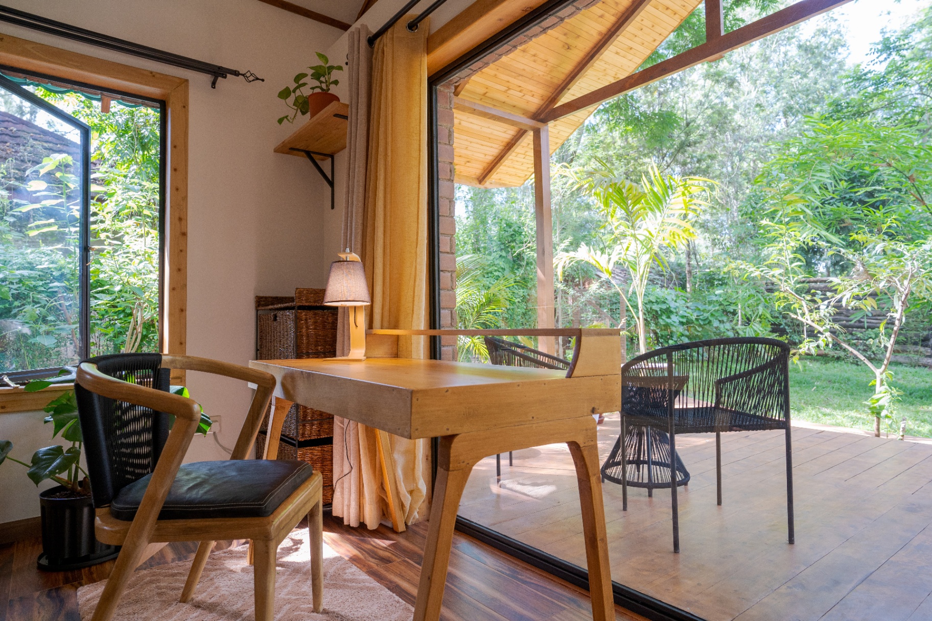 Writing desk in the foreground opening straight onto the deck with woven chairs, side table and a garden bathed in morning sun