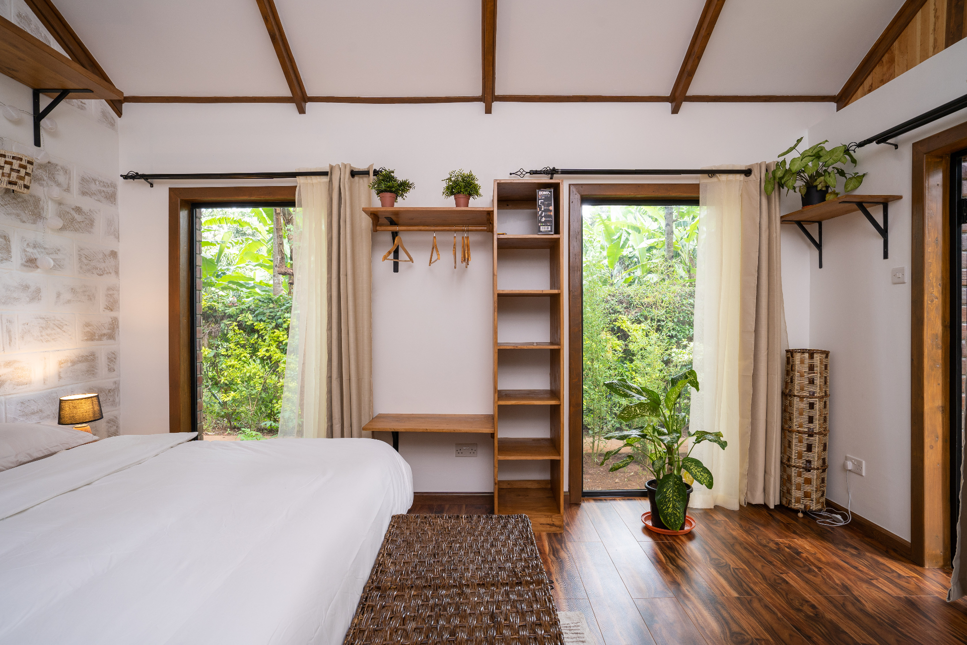 Open wardrobe with wooden hangers between two glass doors that open onto banana palms and dense garden greenery in Cottage 2, with woven textures and potted plants